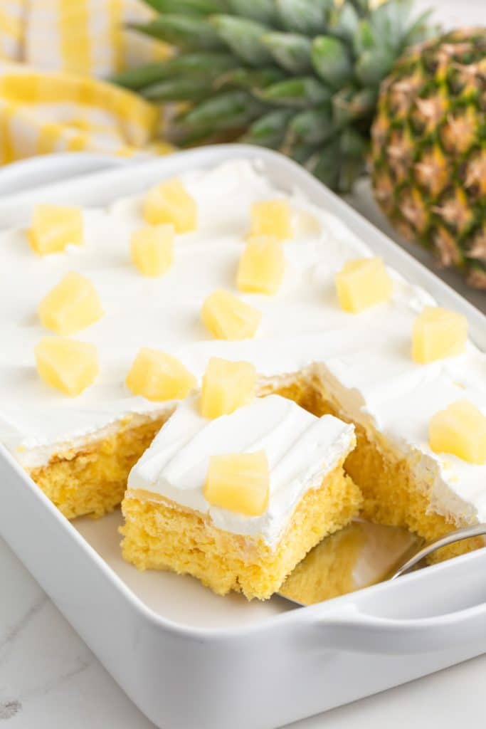 pineapple poke cake in a 9x13 baking pan with one piece on a spatula