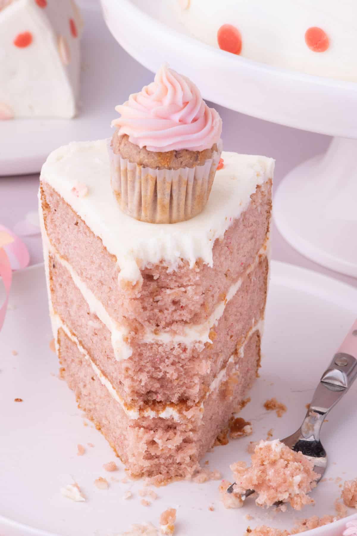 a slice of 3 layer frosted strawberry cake with a mini cupcake on top